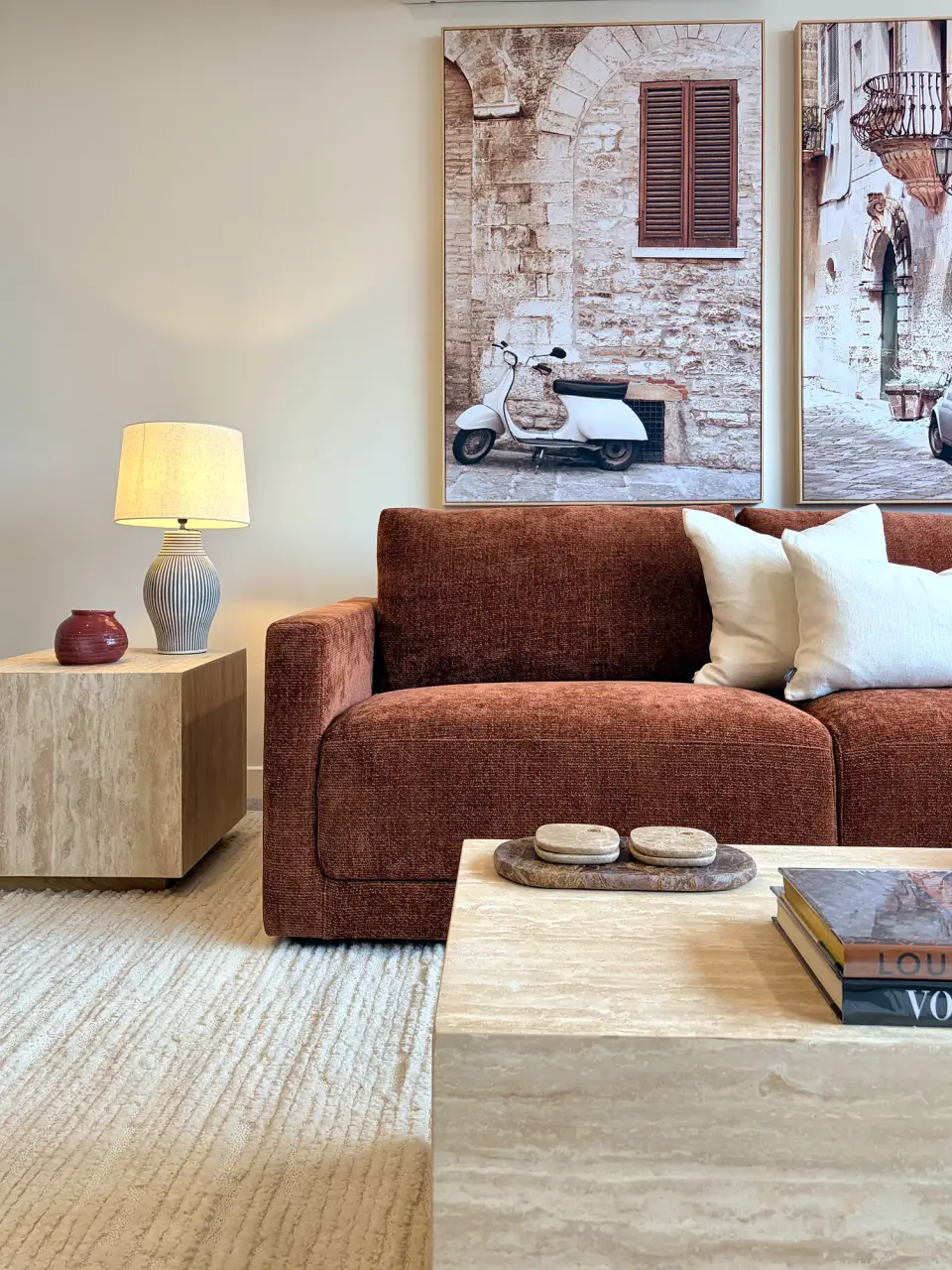 Rust sofa beside a travertine side table and lamp, with framed artwork of a Mediterranean street.