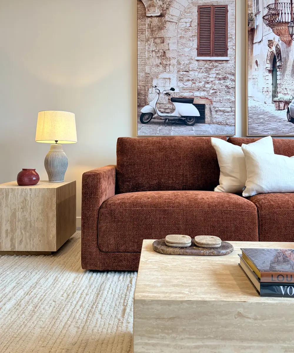 Rust sofa beside a travertine side table and lamp, with framed artwork of a Mediterranean street.