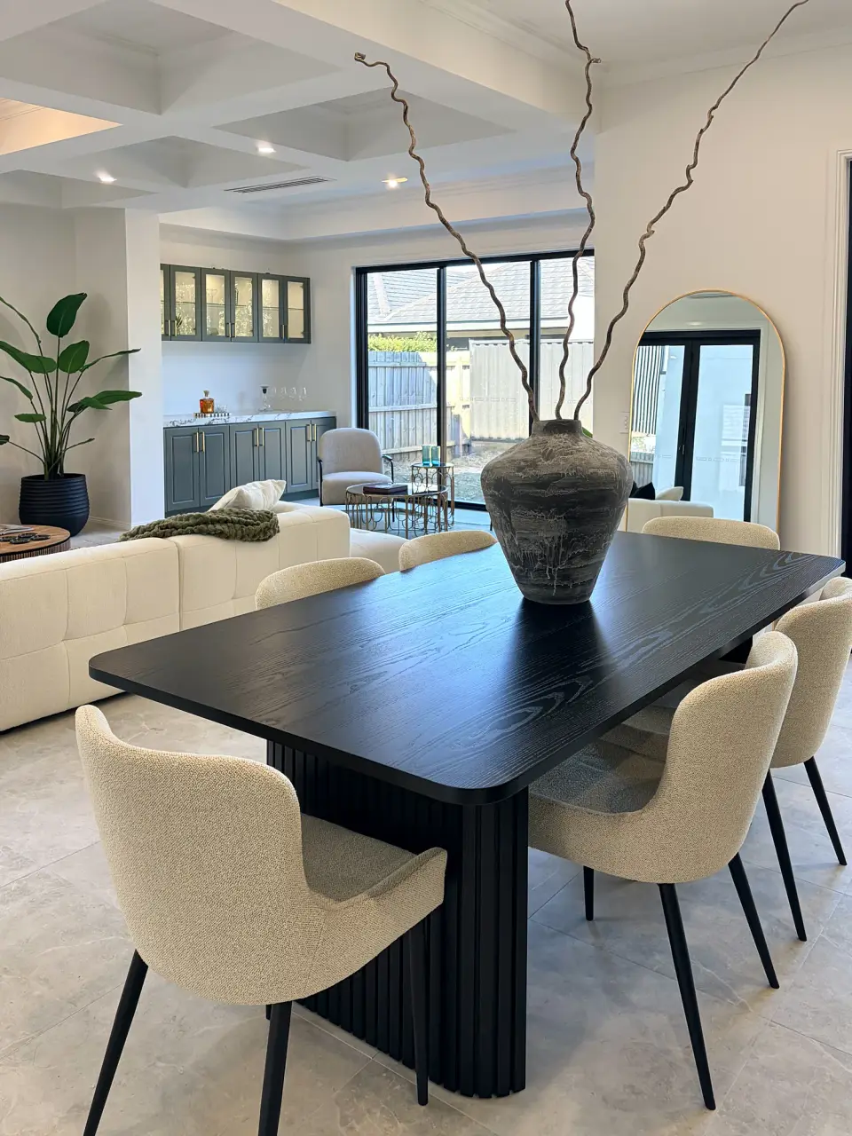 Black dining table with cream upholstered chairs and a tall vase of branches, living area behind.