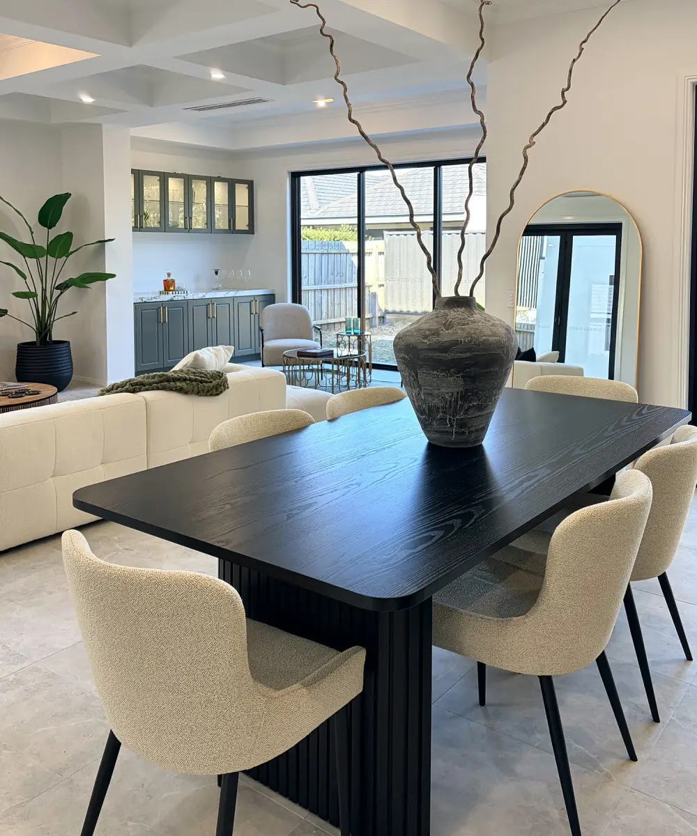 Black dining table with cream upholstered chairs and a tall vase of branches, living area behind.