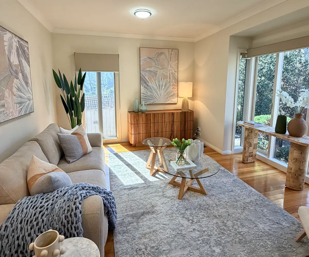 Wide view of the living room with a grey sofa, glass coffee table and rug on timber floors.