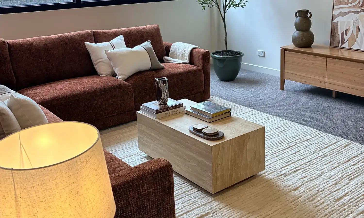Living room seen past a table lamp, with the rust sofa, coffee table and city-view windows.