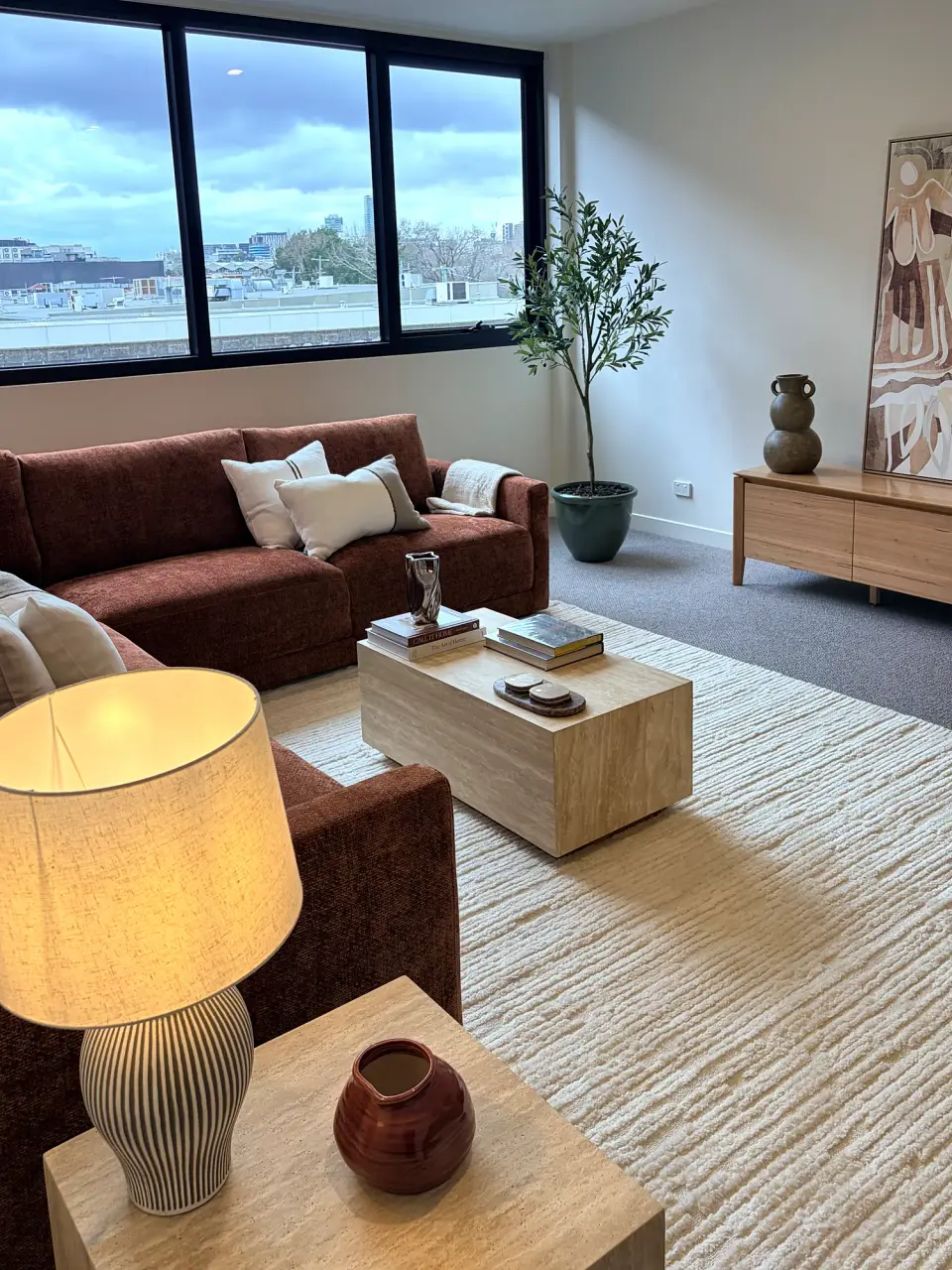 Living room seen past a table lamp, with the rust sofa, coffee table and city-view windows.