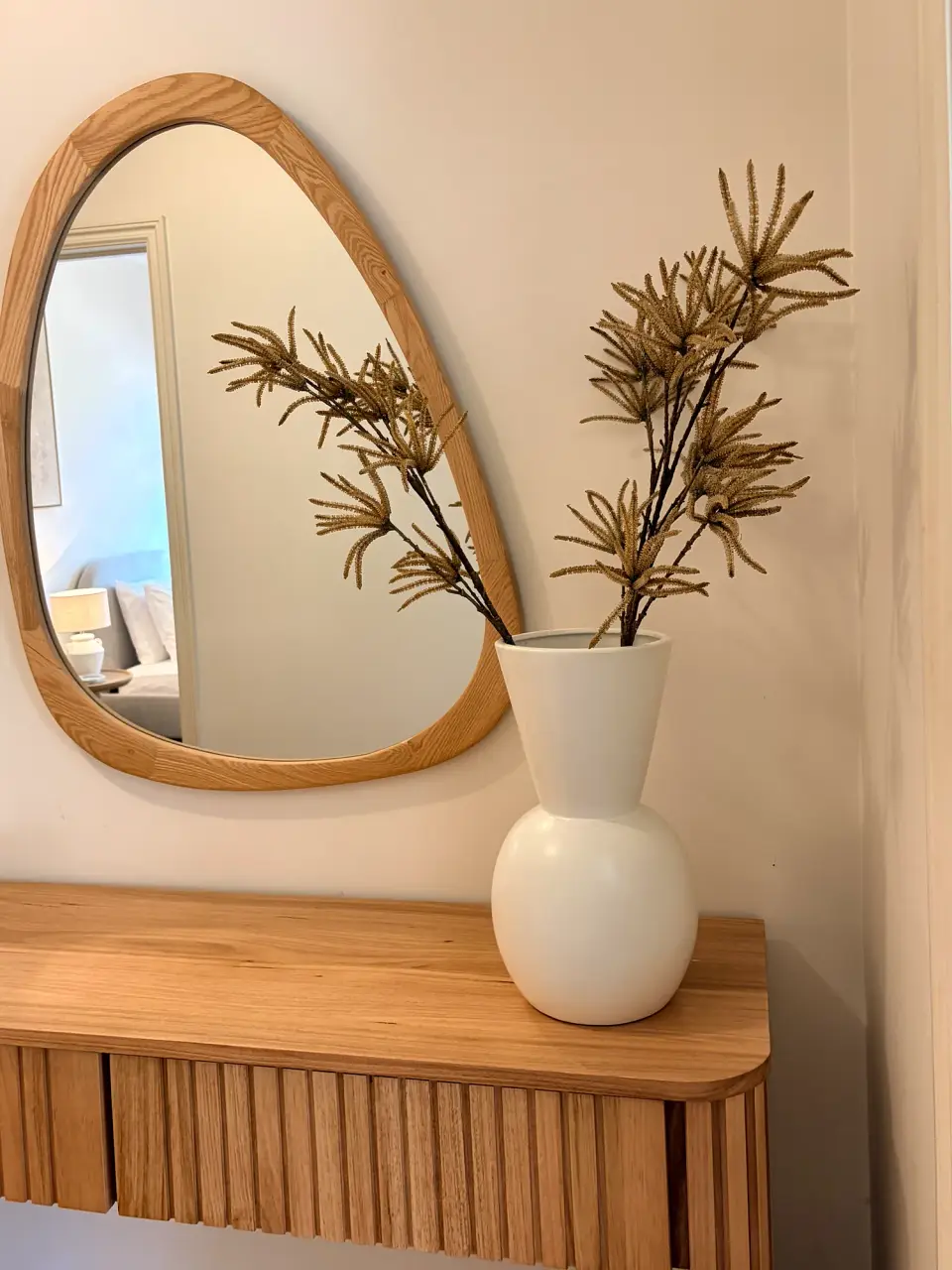 Timber console with an organic-shaped mirror and a white vase of dried foliage.