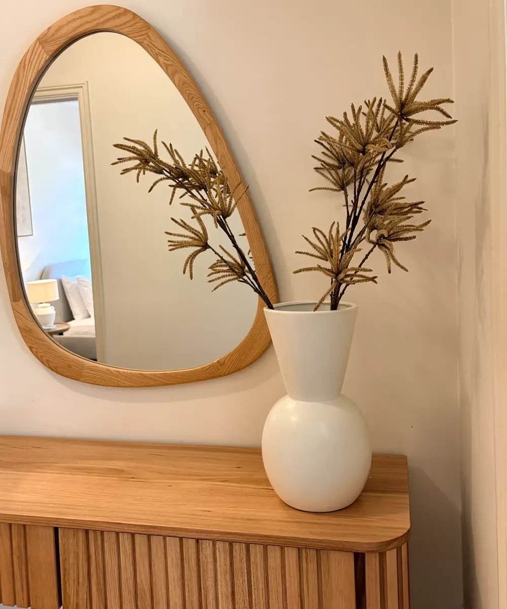 Timber console with an organic-shaped mirror and a white vase of dried foliage.
