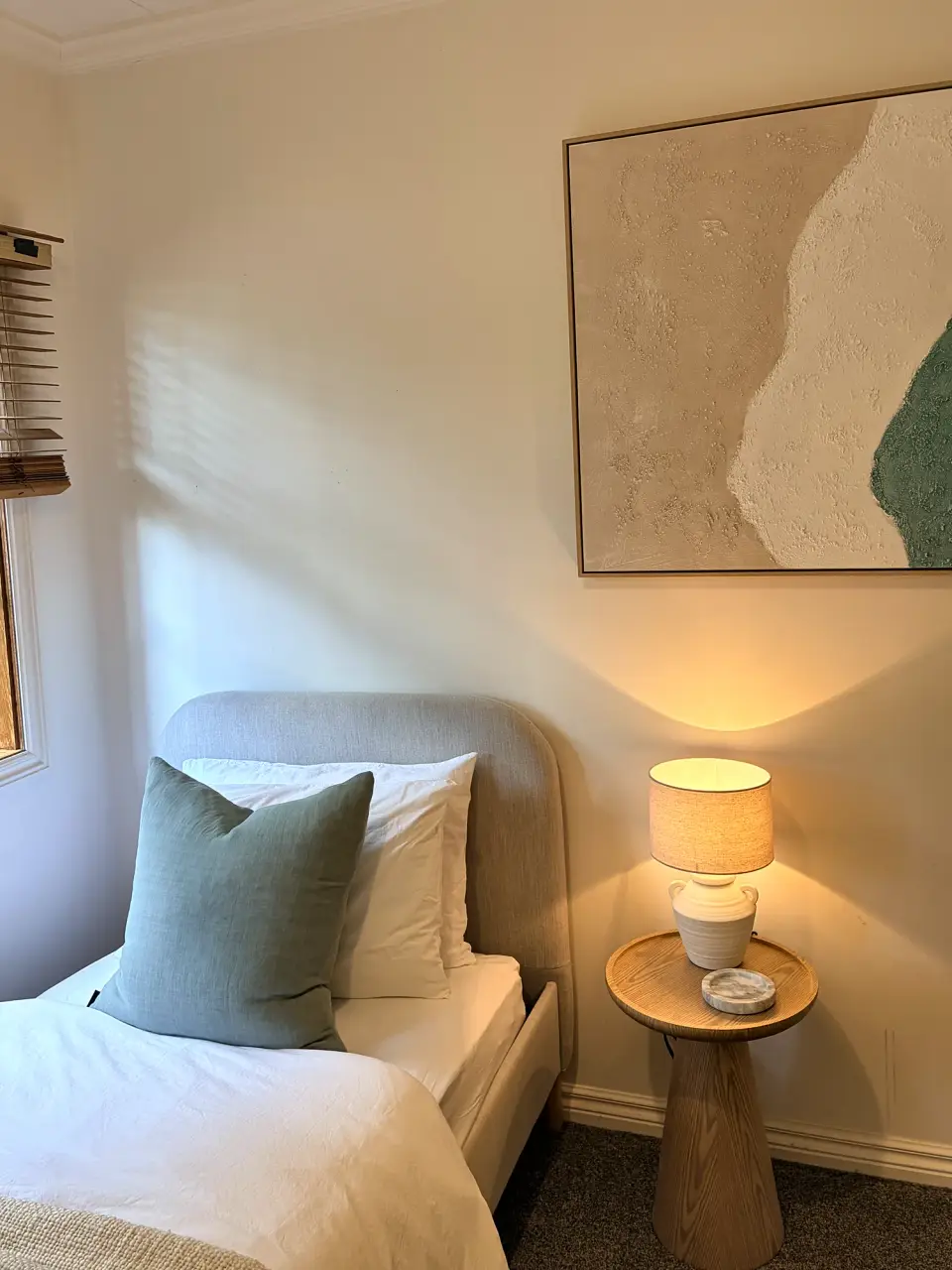 Single bedroom with a sage cushion, round timber bedside table and green abstract artwork.