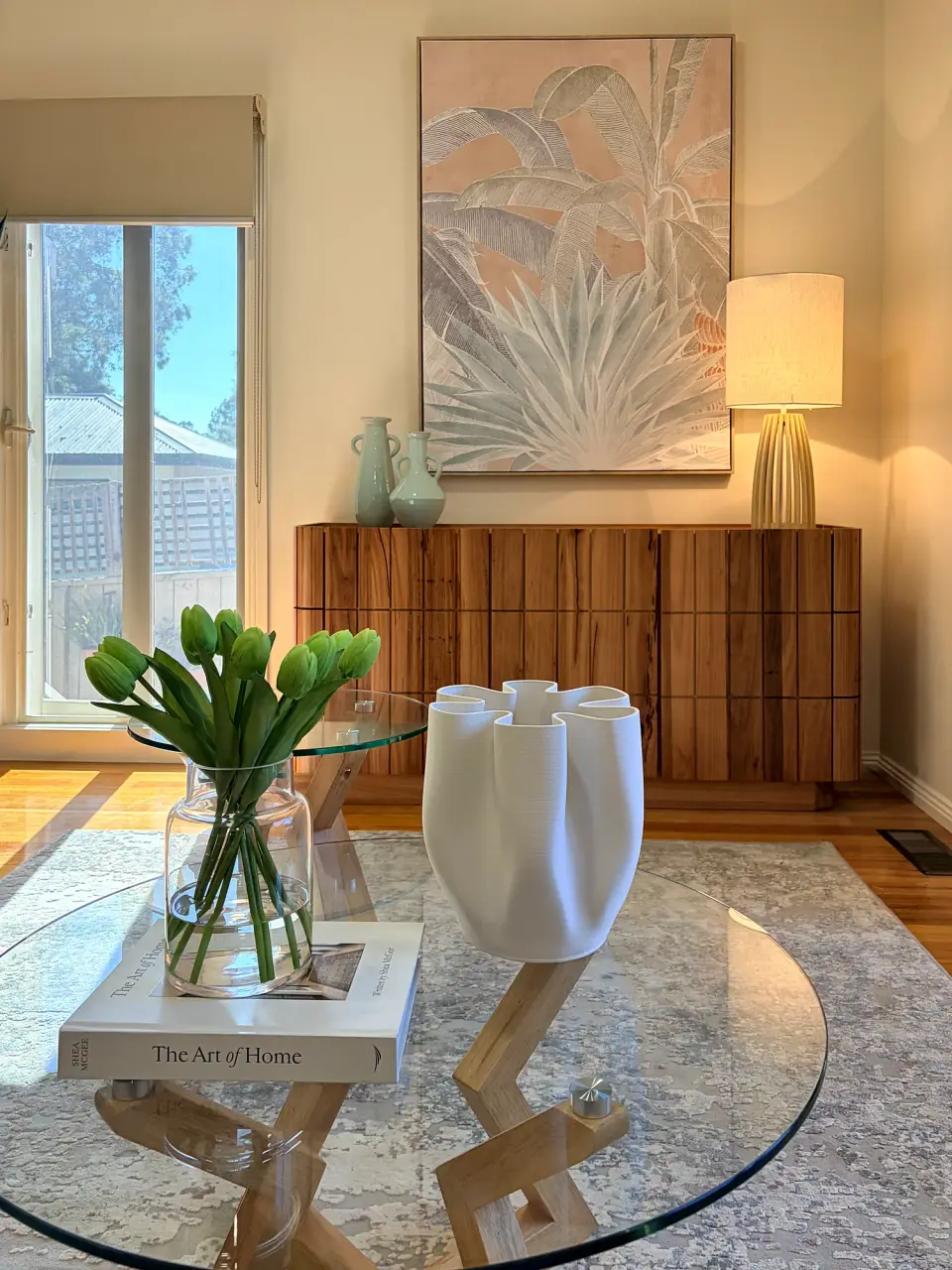 Glass coffee table with tulips and a white sculptural vase, sideboard and artwork behind.