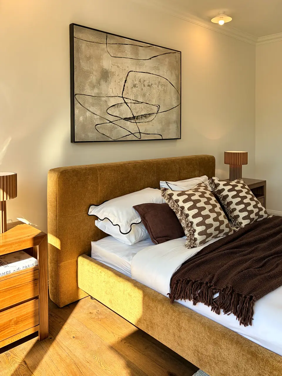 Mustard upholstered bed with patterned cushions and a dark throw in afternoon light.
